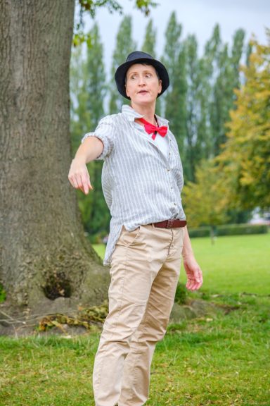 Jowanna Rose Person in a grey shirt and hat gesturing in a park with trees in the background.