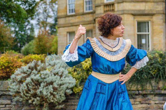 Ali Mylon A woman in a blue vintage dress striking a pose in a garden setting.
